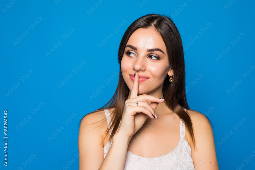 Pretty charming young woman having secret while holding finger on lips and showing silence sign while standing on blue background Pretty charming young woman having secret while holding finger on lips and showing silence sign while standing on blue background
