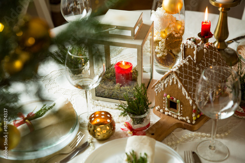 Beautiful served table with decorations, candles and lanterns. Little gingerbread house with glaze on white tablecloth. Living room decorated with lights and Christmas tree. Holiday setting close up