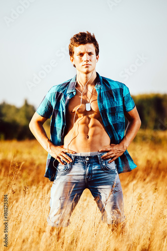 a handsome young sexy man with a strong muscular torso in an unbuttoned shirt is standing on a meadow in nature outside city