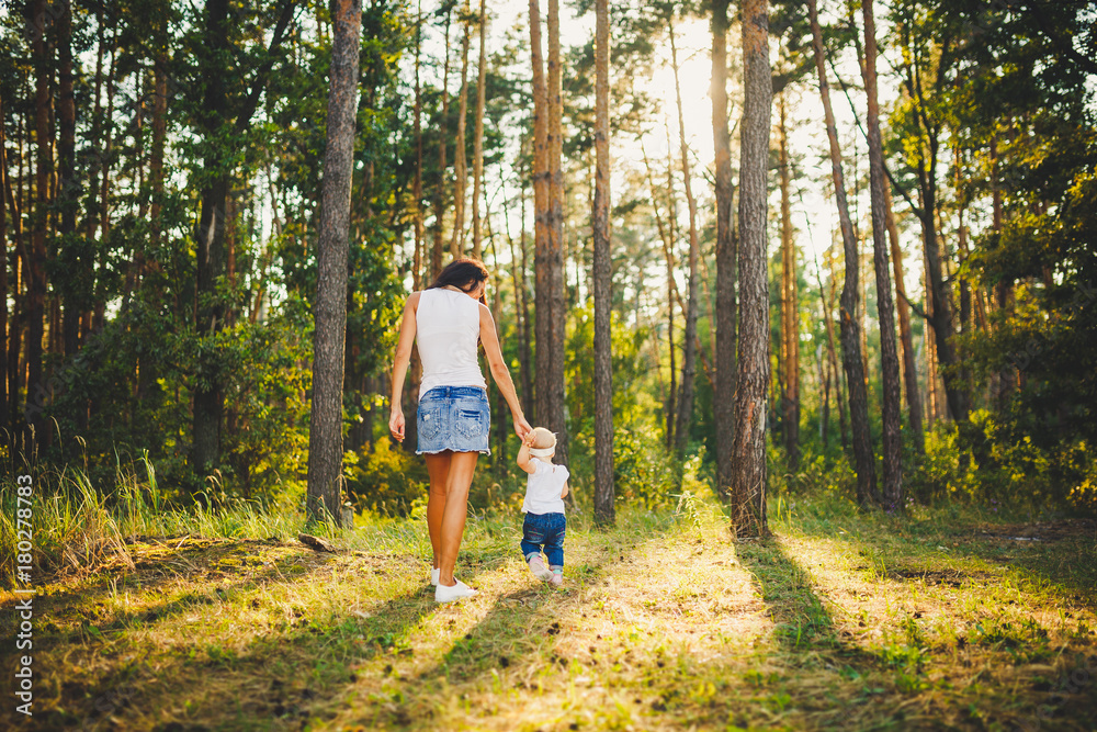 Obraz premium beautiful and stylish mother leads the hand of a girl of one year from birth in the park against the background of tall coniferous trees. Family values mom learns to walk child