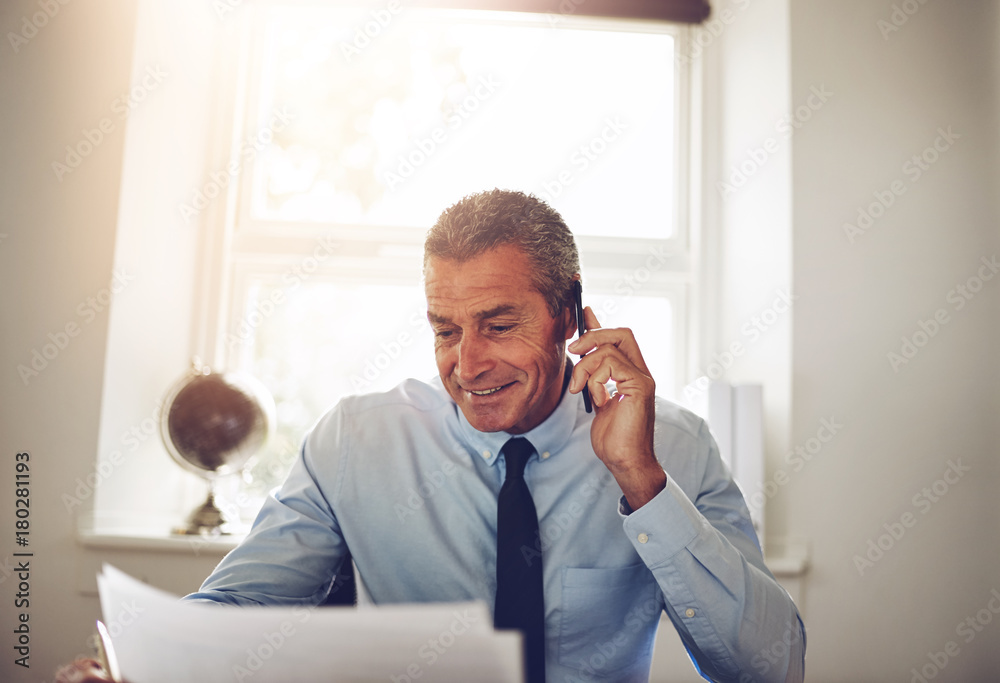 Smiling businessman discussing paperwork over the phone in his o Smiling businessman discussing paperwork over the phone in his o