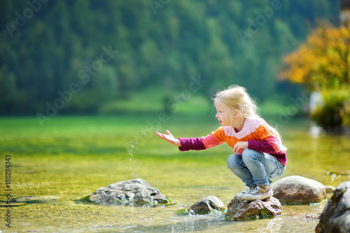 Adorable girl playing by Konigssee lake in Germany on warm summer day. Cute child having fun feeding ducks and throwing stones into the lake.