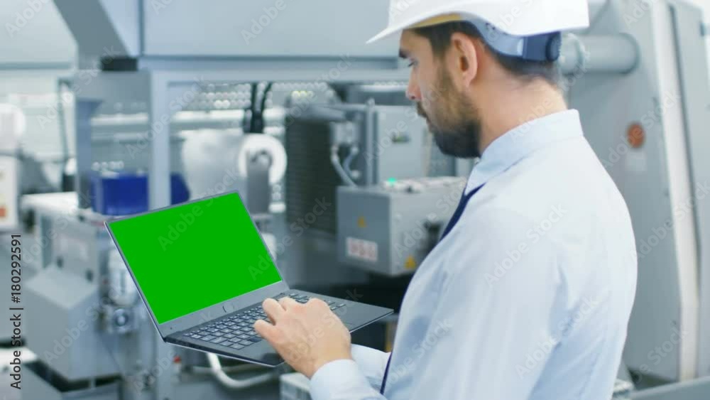 Industrial Engineer in Hard Hat Holds Laptop while Working on 3D Turbine/ Engine Component in CAD Program. He's Standing in the Middle of Modern Electronics Manufacturing Factory. 