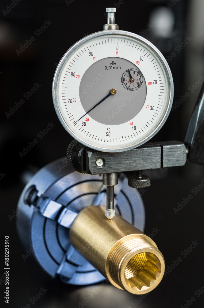 Measurement of permissible tolerance of the brass workpiece clamped in the chuck. Gold fitting and dial indicator in holder on black background. Concept for engineering, education and industry.