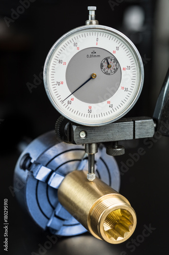 Measurement of permissible tolerance of the brass workpiece clamped in the chuck. Gold fitting and dial indicator in holder on black background. Concept for engineering, education and industry.