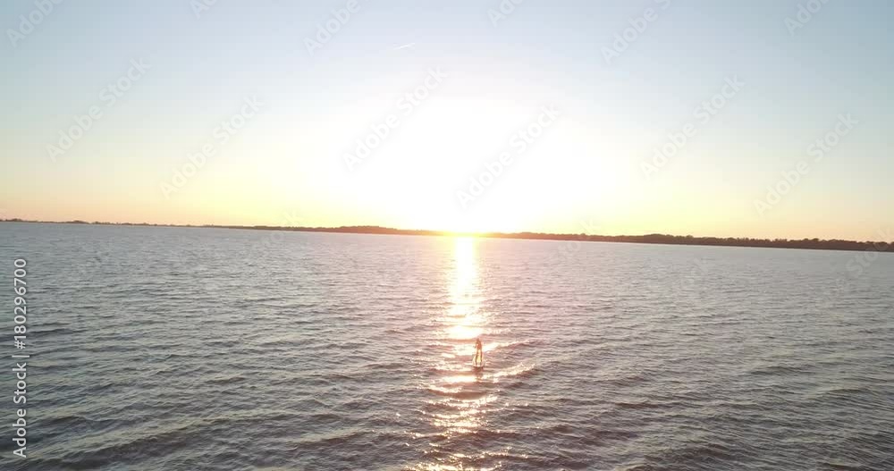 A far shot of a woman paddle boarding away from the sunrise A far shot of a woman paddle boarding away from the sunrise