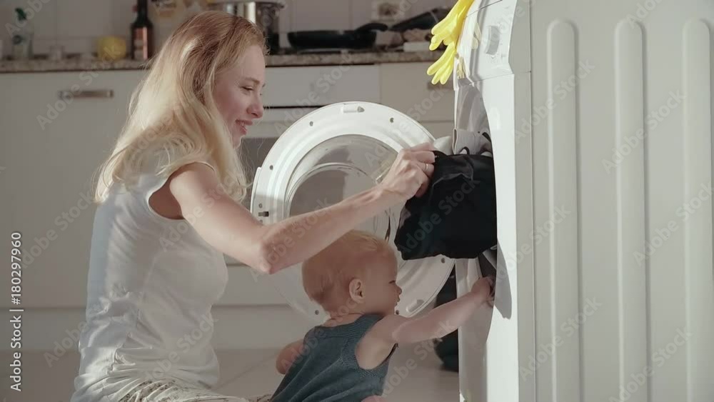 Young mother with baby sitting on floor and putting laundry in washing machine