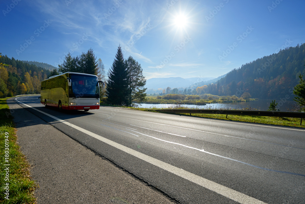 Glowing sun over the landscape with a bus traveling on the road around a lake and forested mountains in autumn