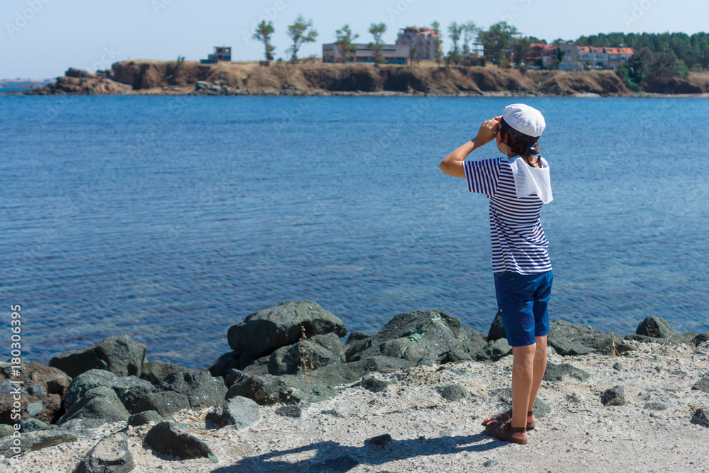 A boy in a striped T-shirt and a cap on the seashore and looks through binoculars.