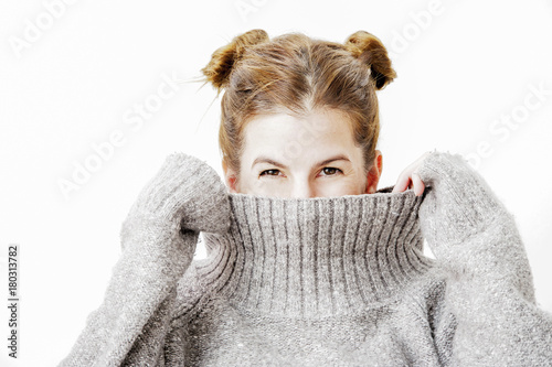 Obraz na plátně Funny cute young woman is smiling into the camera while covering her face with the collar of her sweater