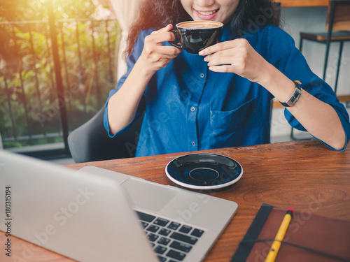 Young asian woman sitting in coffee shop at wooden table, hand holding coffee cup, Using laptop browsing internet, chatting, blogging, shopping onlie. photo