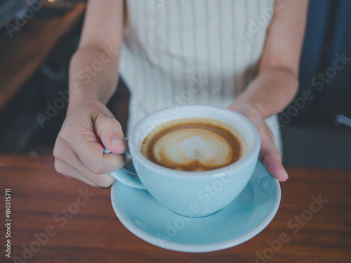 Young asian woman barista with a smile, holding a glass cup of coffee at bar counter.