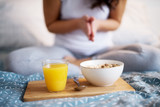 Close up of breakfast and juice on a wood saucer on the bed while the woman behind sitting and prying.