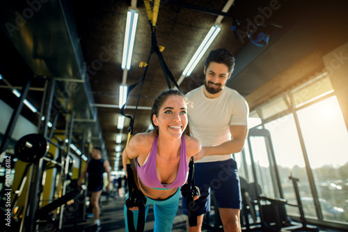Beautiful slim happy girl exercising in front of the smiling personal trainer in the gym.