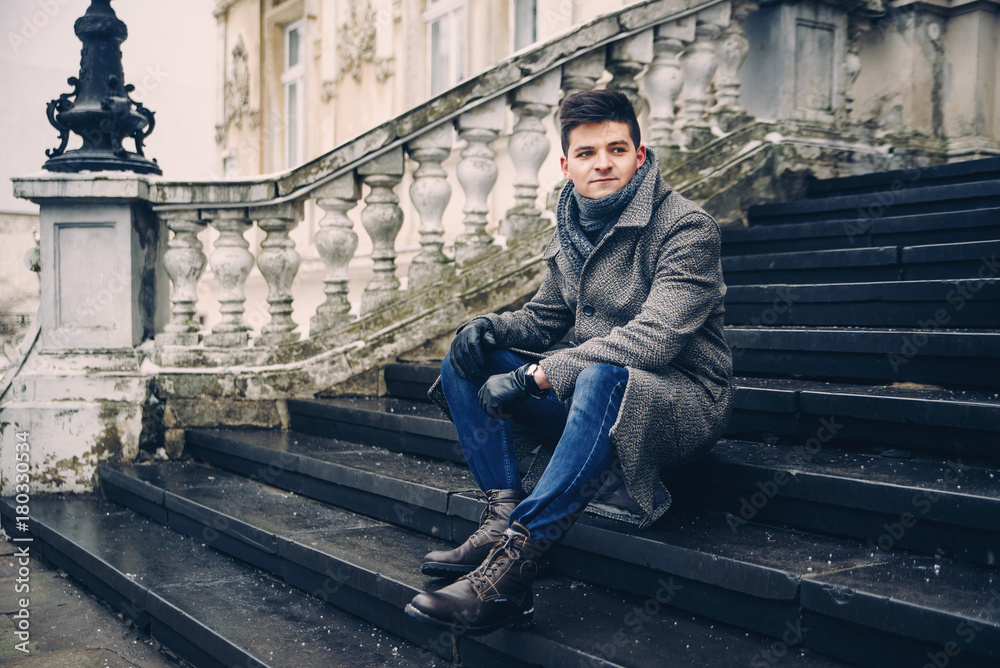 Young stylish man in warm gray coat and leather gloves sitting on the stairs.