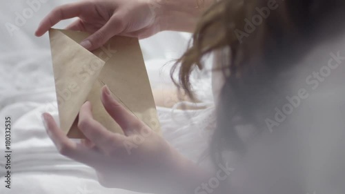Top view of unrecognizable woman with long hair sitting in bed on early morning and opening envelope with letter from lover photo