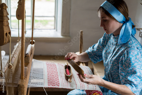 The woman is weaving the colorful cotton robe or dress by using wooden loom in the local village in Russia