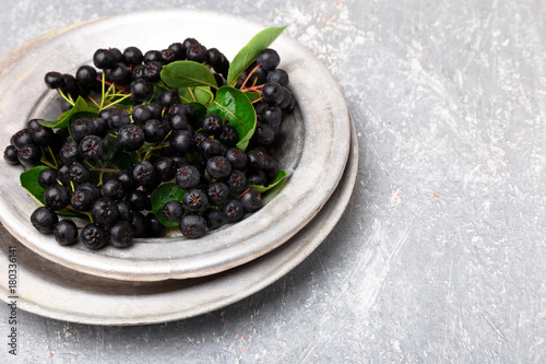 Chokeberry in silver metal bowl on grey background. Aronia berry with leaf. Top view. Copy space.