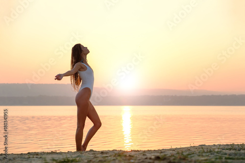 Young woman in strings swimsuit stands on the beach in sunrise 