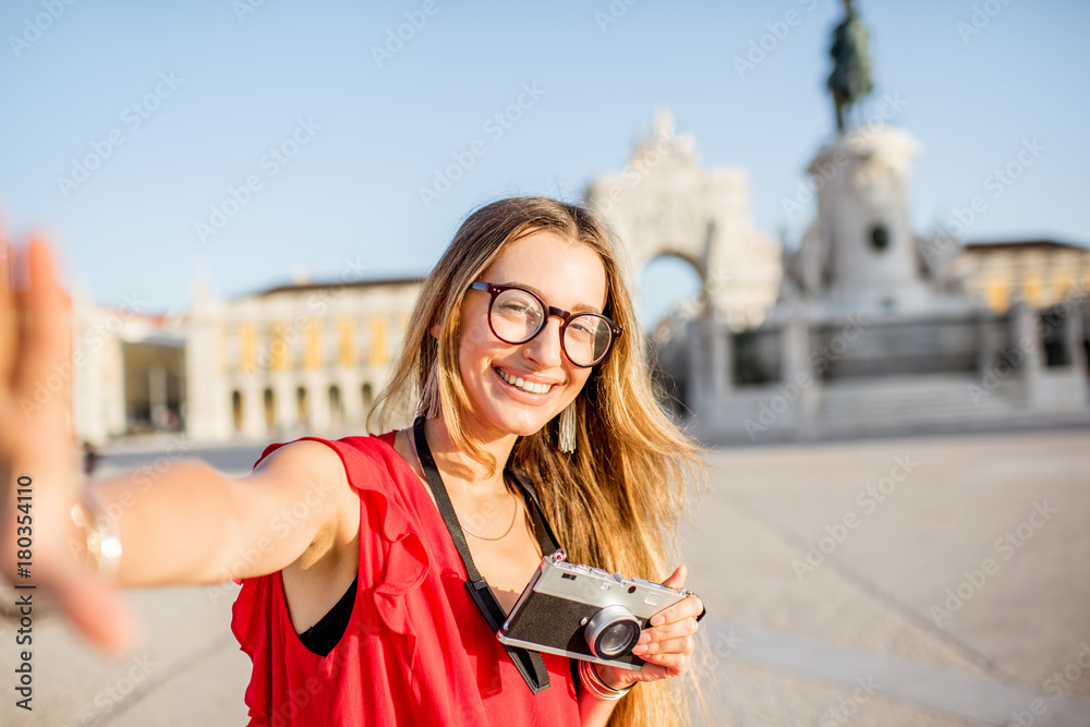 Fototapeta premium Lifestyle portrait of a young woman tourist standing on the main square with statue and triumphal arch on the background during the morning light in Lisbon city, Portugal