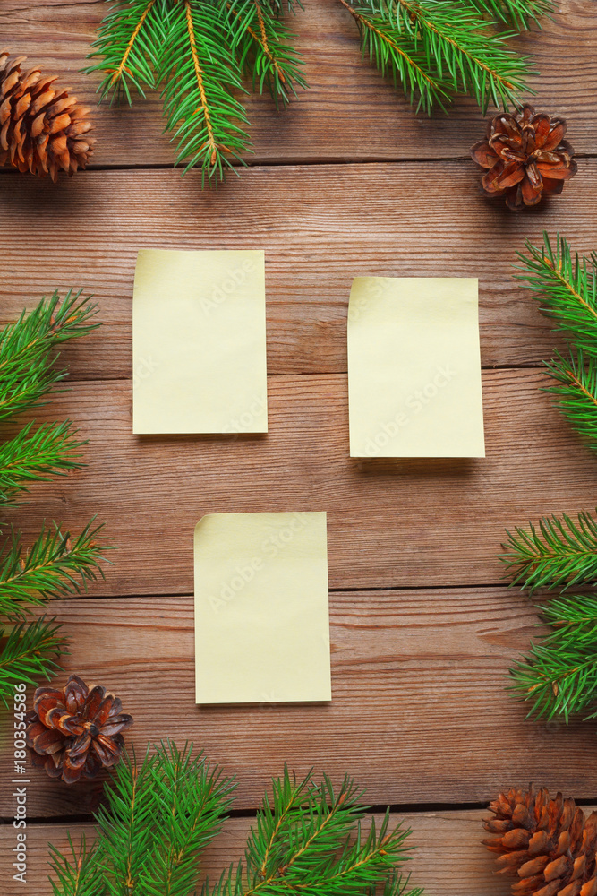 Fototapeta premium Christmas frame with stickers for notes, with spruce branches and cones on a natural wooden background.