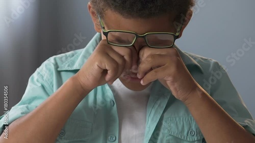Little biracial boy in glasses doing homework, rubbing eyes, strained eyesight
