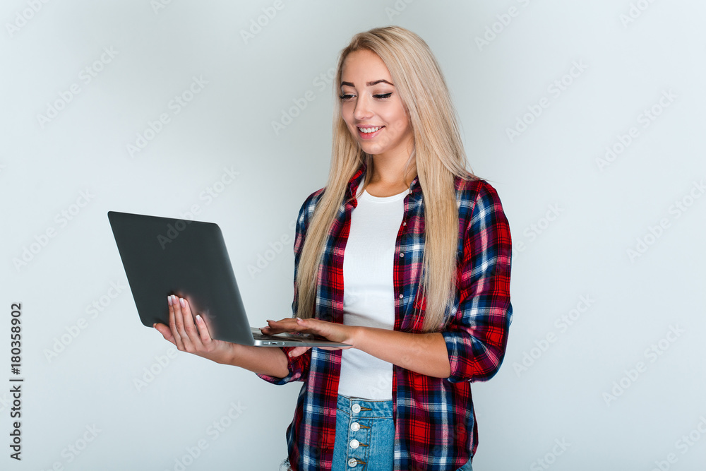 Studying is fun. Beautiful young woman with blond hair using her laptop with smile while standing against grey background Studying is fun. Beautiful young woman with blond hair using her laptop with smile while standing against grey background