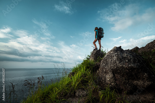 Lady hiker with backpack standing on the stone and enjoying sea view
