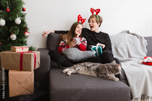 Portrait of young beautiful couple sitting on sofa at home and wearing deer horns while happily spending time together with cat near