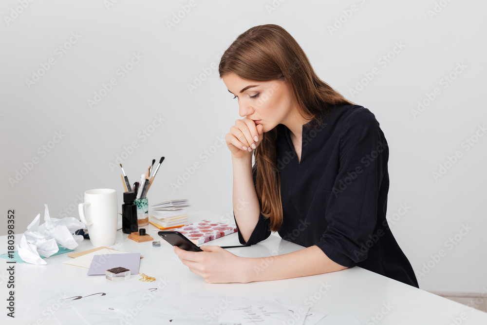 Portrait of young beautiful lady sitting at the white desk and thoughtfully looking in her cellphone isolated Portrait of young beautiful lady sitting at the white desk and thoughtfully looking in her cellphone isolated