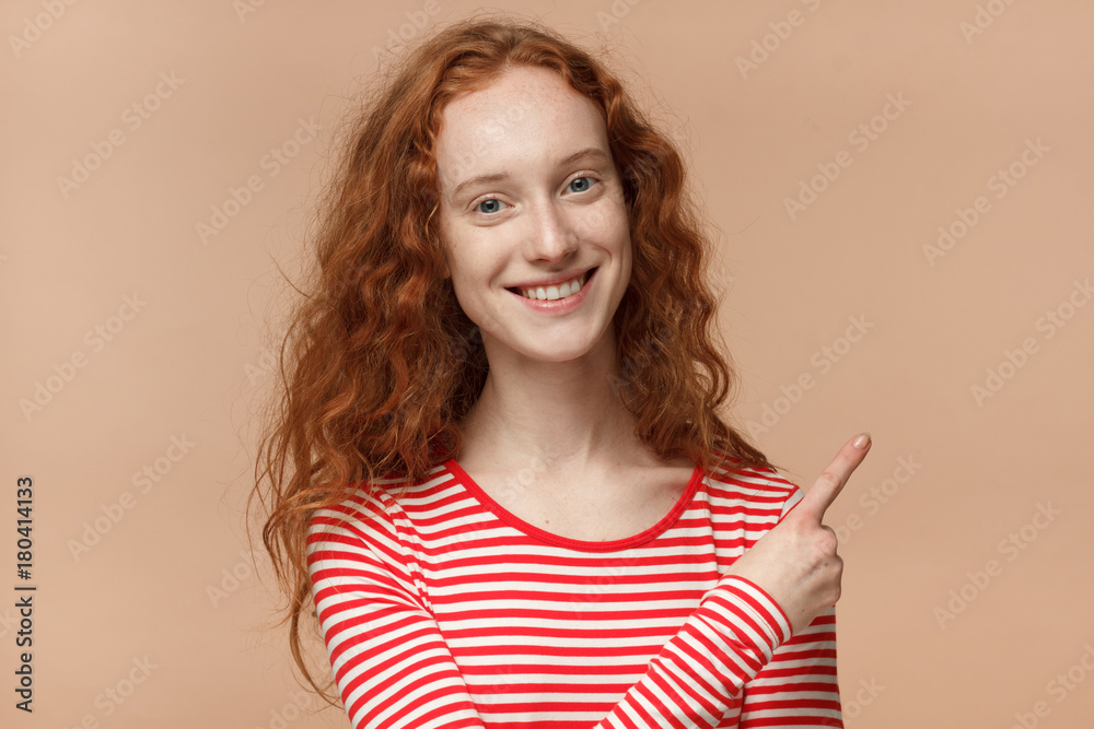 Indoor closeup of young beautiful European woman with red loose curly hair isolated on peach background pointing to important information on right side of picture smiling happily and looking calm Indoor closeup of young beautiful European woman with red loose curly hair isolated on peach background pointing to important information on right side of picture smiling happily and looking calm