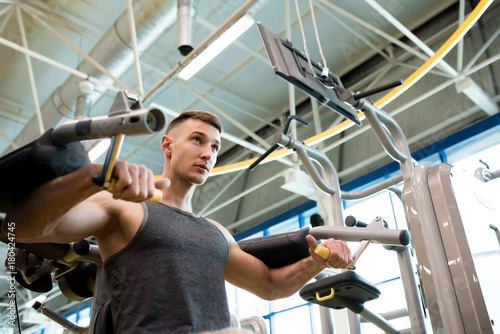 Low angle portrait of handsome young man pumping arm muscles using machines in gym, copy space
