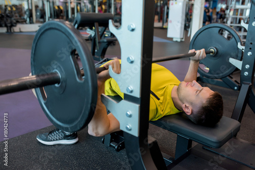 Portrait of young man doing weightlifting exercises with heavy barbell on bench during workout in modern gym