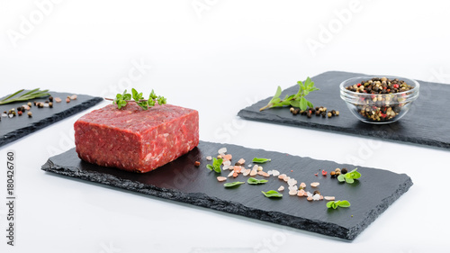 Three slate boards with piece of raw ground beef, glass bowl with whole pepper, pink salt and fresh marjoram and rosemary leaves photo