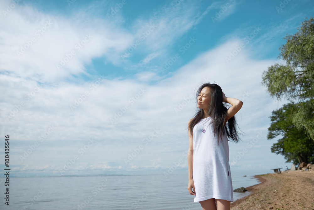 Ecstatic young Asian brunette woman enjoying vacations by the sea, closing eyes and smiling joyfully, feeling happy and carefree standing against blue sky background with copy space for your content Ecstatic young Asian brunette woman enjoying vacations by the sea, closing eyes and smiling joyfully, feeling happy and carefree standing against blue sky background with copy space for your content
