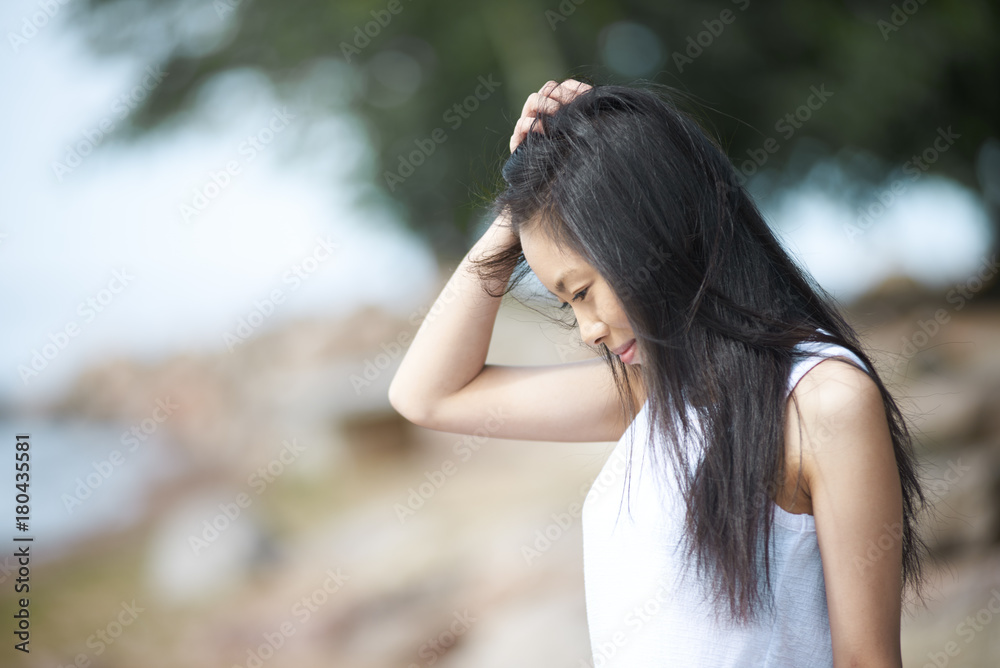 Sideview portrait of charming Chinese young woman wearing white top posing outdoors, looking down and touching her long black hair, smiling mysteriously, enjoying nice summer day, Blurred background Sideview portrait of charming Chinese young woman wearing white top posing outdoors, looking down and touching her long black hair, smiling mysteriously, enjoying nice summer day, Blurred background