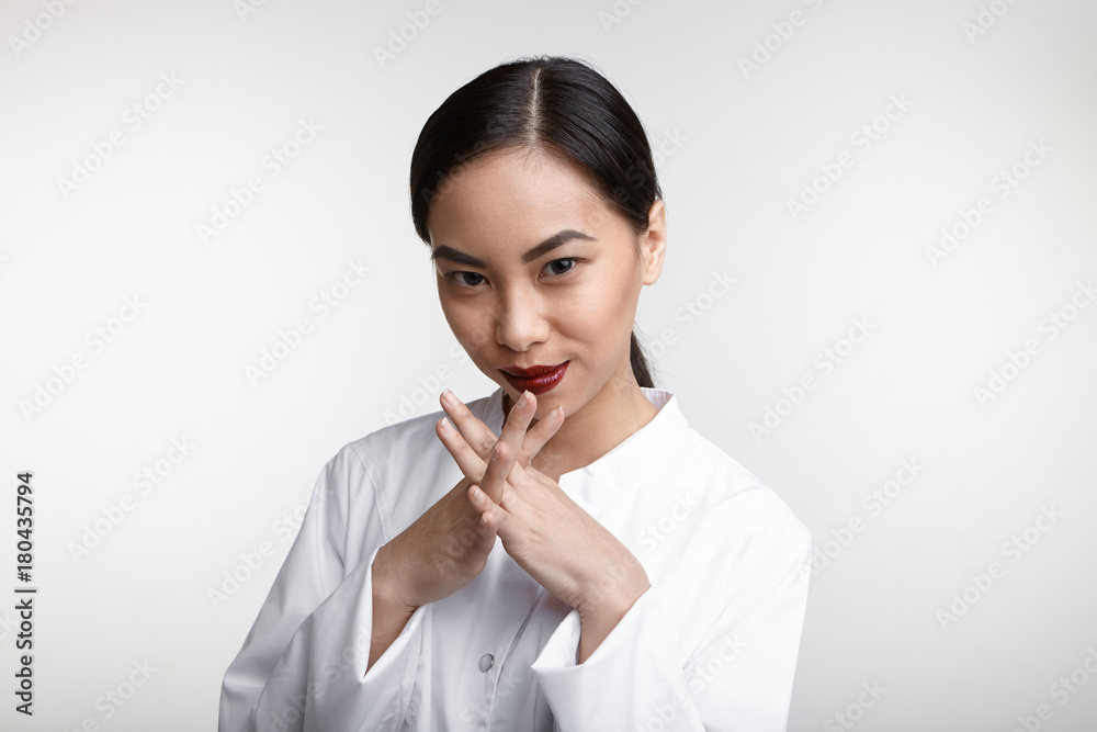 Horizontal shot of attractive successful young Korean woman plastic surgeon with bright make up looking at camera and smiling mysteriously, clasping her hands, ready to see her next patient Horizontal shot of attractive successful young Korean woman plastic surgeon with bright make up looking at camera and smiling mysteriously, clasping her hands, ready to see her next patient
