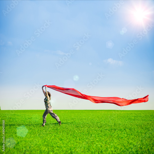 Beautiful young woman jumping on a green meadow with a colored red tissue. Happy female in wheat field with fabric. Summer picnic and freedom concept.