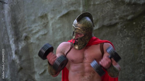 Horizontal shot of a handsome bearded mature medieval warrior in a helmet and red cape working out with heavy weights flexing his muscles outdoors strengthen powerful energetic.