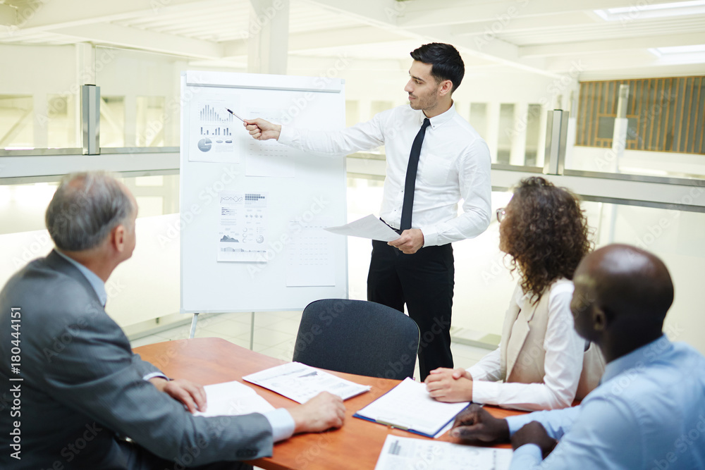 Young confident speaker pointing at financial data on whiteboard and explaining chart Young confident speaker pointing at financial data on whiteboard and explaining chart