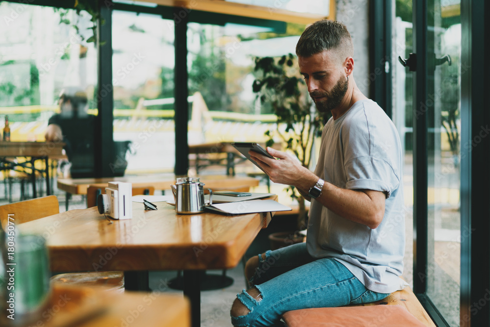 Handsome bearded student preparing coursework project while browsing information in the Internet and writing down general concepts. Young hipster guy holding tablet while sitting in modern coffee shop