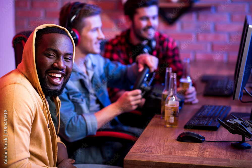 Portrait of positive stylish african young man is sitting at table with computer and looking at camera with joy. Cheerful cute gamers with headphones are playing video game in background. Copy space