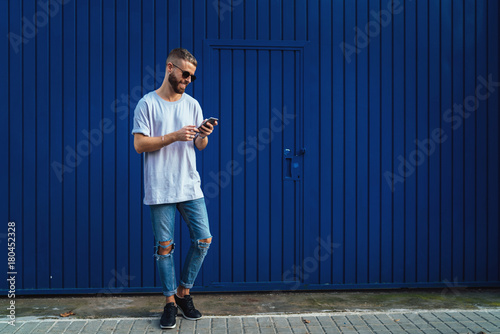 Smiling student guy chatting online via social network on a mobile phone while walking the street. Stylish hipster male checking email box on a smartphone while standing on a blue wall background.