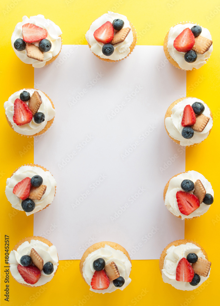 Obraz premium Cupcakes with cream, strawberry, blueberry and cookie on yellow background in the form of a frame. Image with copy space for the confectionery menu, cards, greetings, birthday invitations. Top view