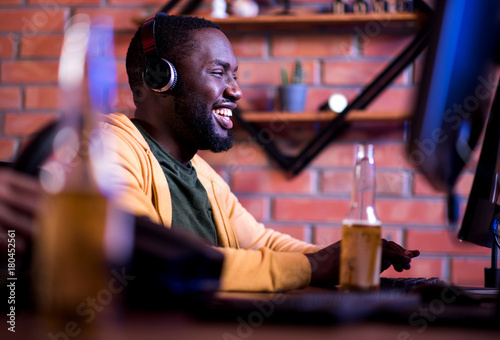 Lets win. Profile of joyful african gamer in headphones is playing on computer while sitting at table with beer and expressing gladness. He is spending time at home with pleasure