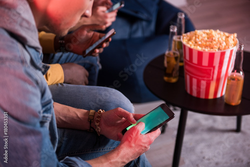 Useful gadget. Close-up of screen of mobile phone in hands of young man who is sitting on couch. He is sending message while resting with friends at home. Bottles of beer and popcorn are in background