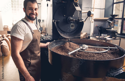 Portrait of happy unshaven male having job at factory. He controlling preparing coffee beans in roaster equipment. Occupation concept