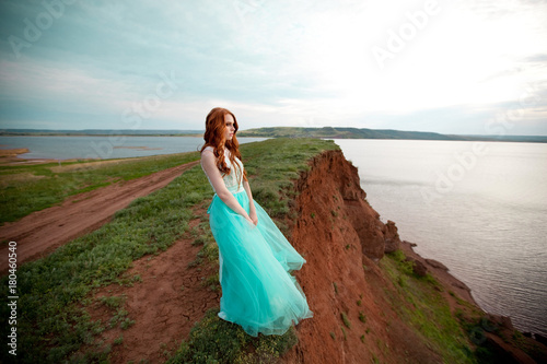 beautiful young girl with red hair in a lavish dress walking in the nature near the lake