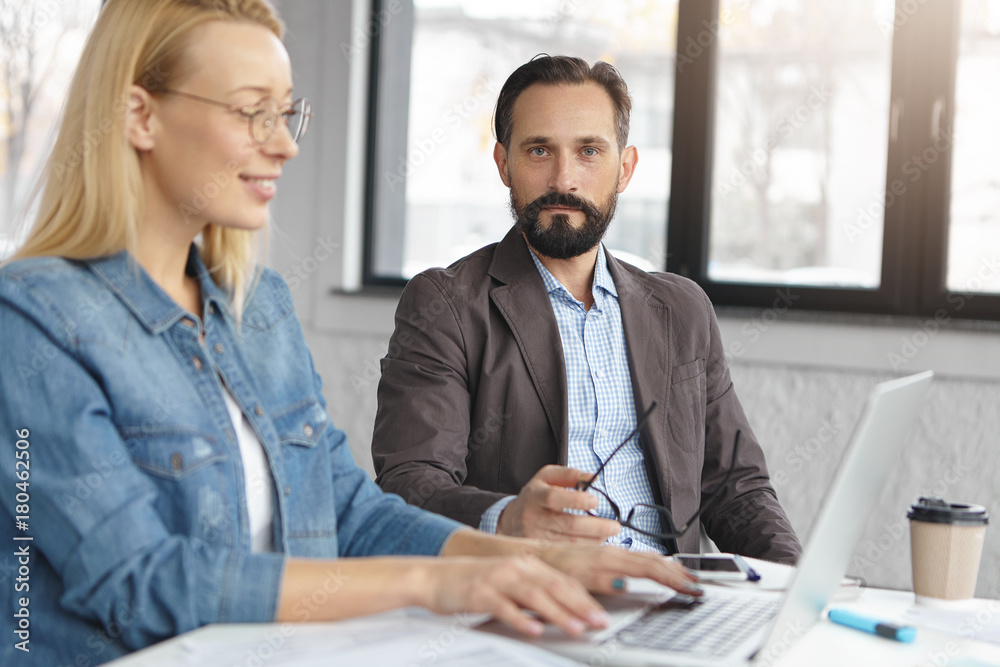 © wayhome.studio - Serious bearded man in jacket and shirt controls female`s work, collaborate together, work on common business presentation on generic laptop computer, sit over office interior. Teamwork concept
