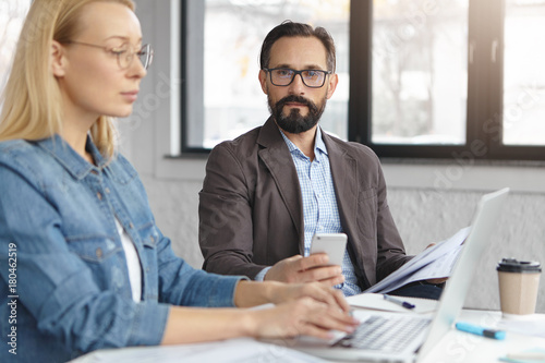 Canvastavla Middle aged bearded male manager dressed formally dictates information to his female secretary who sits near him and keyboards on laptop, have good business ties and relationships